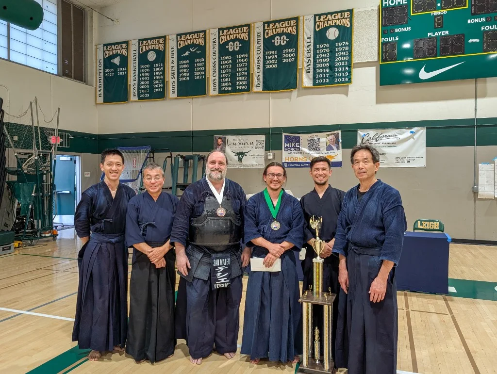 Six kendo practitioners in keikogi and hakama posing in a high school gym after the 42nd NCKF Championships, with a medal on a green ribbon and a trophy